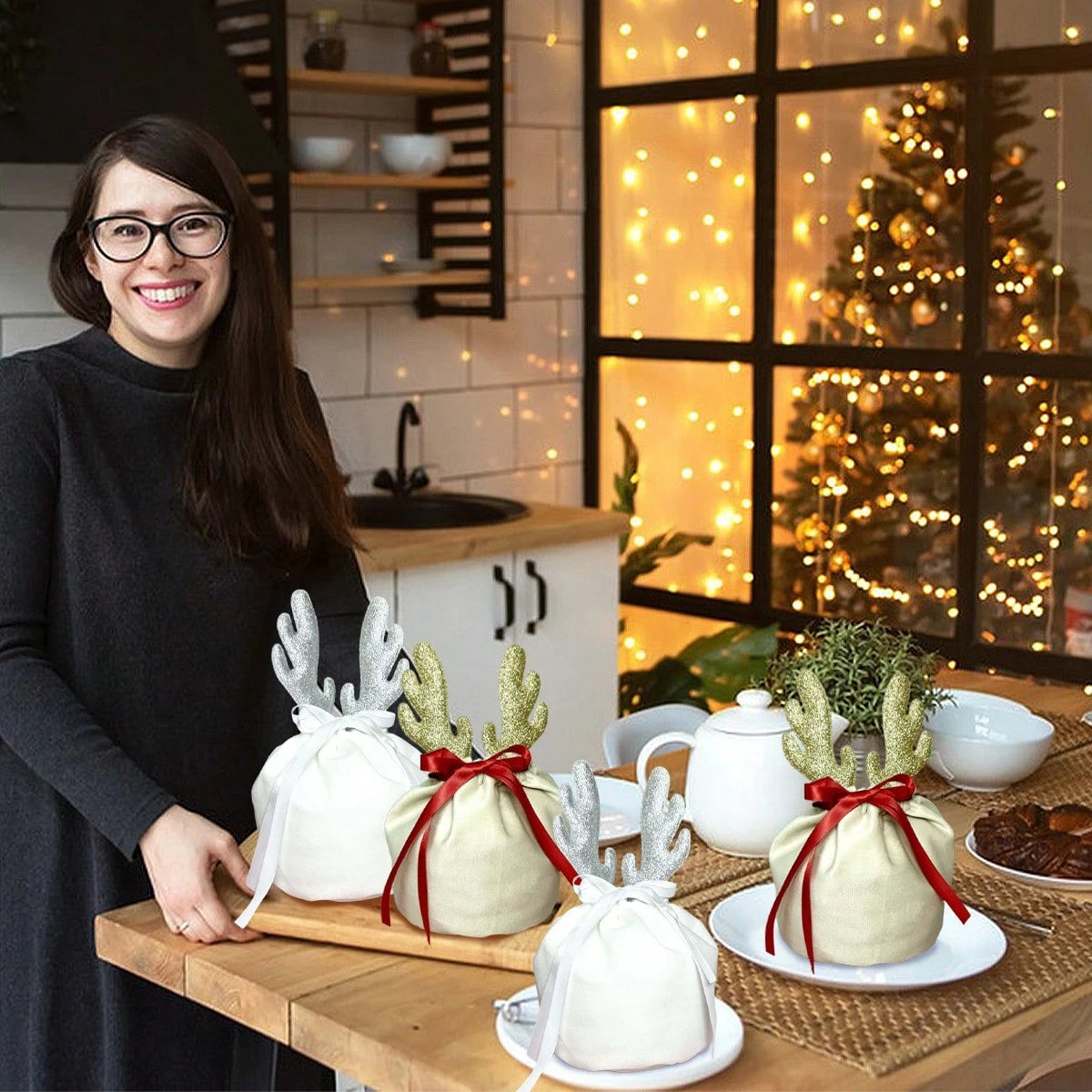 Smiling woman with glasses displaying Christmas velvet drawstring gift pouches with glitter antlers in a cozy kitchen