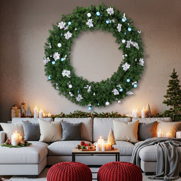 Large green Christmas wreath with white flowers and blue ornaments above cozy sofa with candles and red poufs