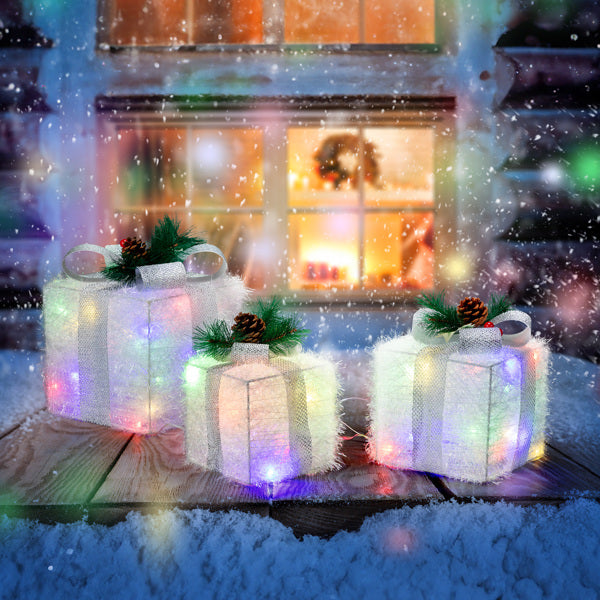 Three multicolor LED lighted Christmas gift boxes with ribbons and pine decorations on snowy wooden surface