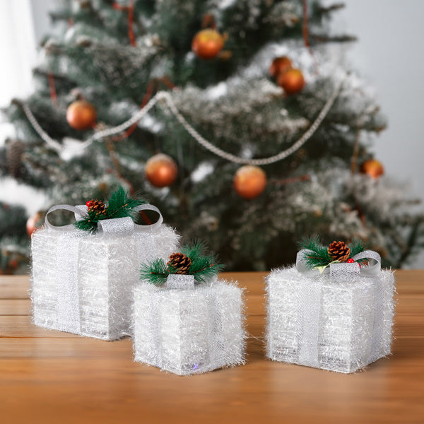 Set of 3 white lighted Christmas gift boxes with silver ribbons and pinecone accents on wooden table in front of decorated Christmas tree