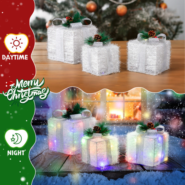 Set of 3 white LED lighted Christmas gift box decorations with pine cones on wooden table, daytime and nighttime views
