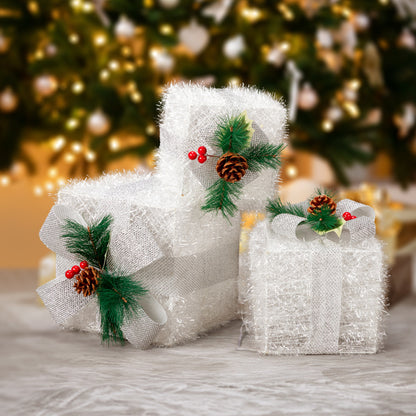 Set of three white lighted Christmas gift boxes decorated with pine cones, green pine sprigs, and red berries on a marble surface