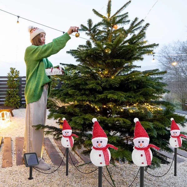 Woman in green sweater decorating outdoor Christmas tree with warm white lights, surrounded by snowman stake lights