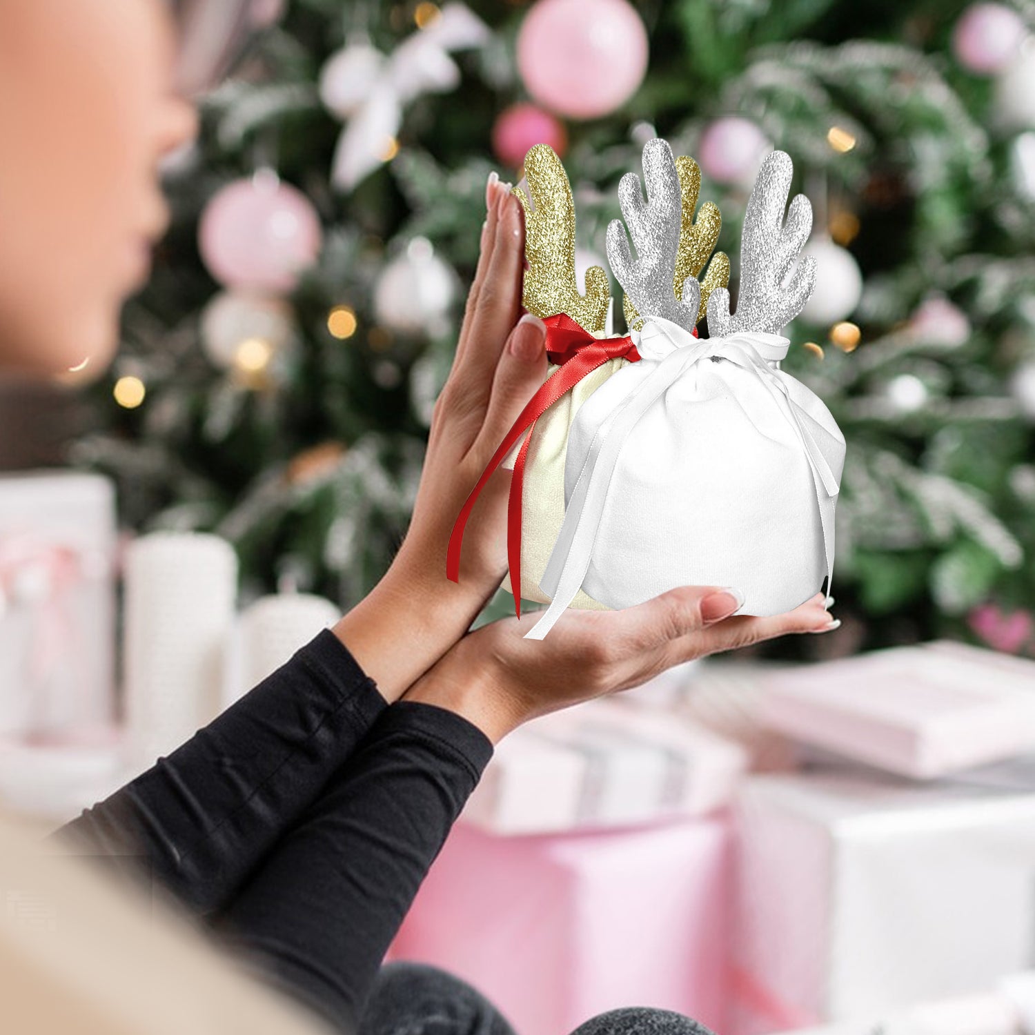 Hands holding white and khaki Christmas gift bags with glitter reindeer antlers near decorated Christmas tree