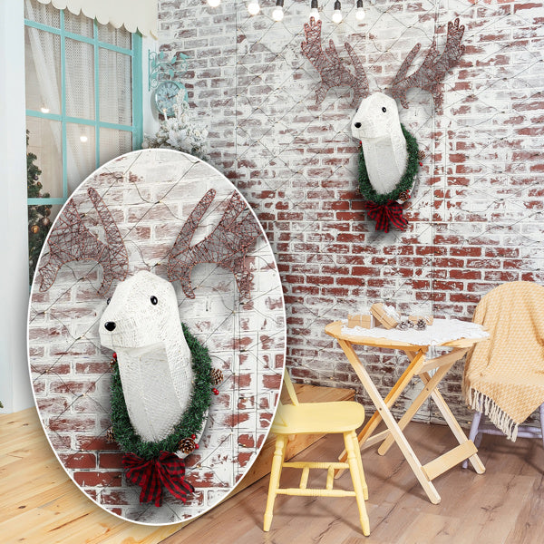 Indoor Christmas decor featuring a warm white LED lighted reindeer head with a green wreath and red bow on a white brick wall