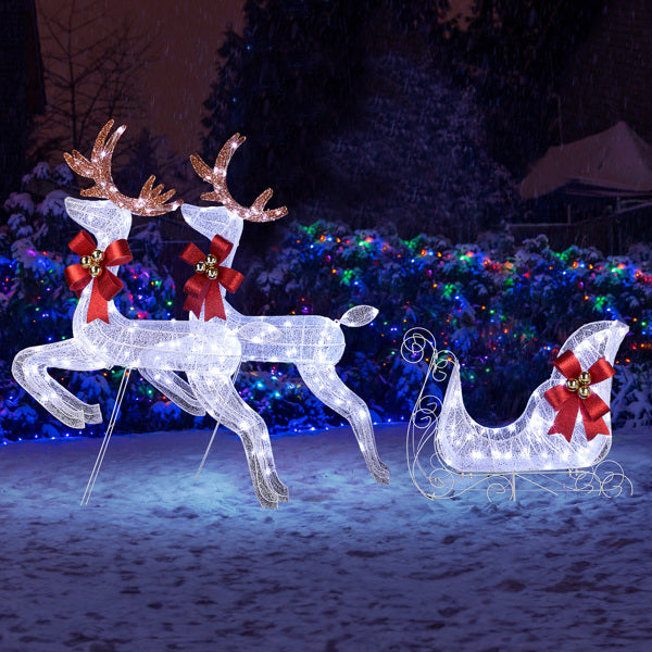 White LED light-up Christmas reindeer and sleigh decorations with red bows and jingle bells in snowy yard